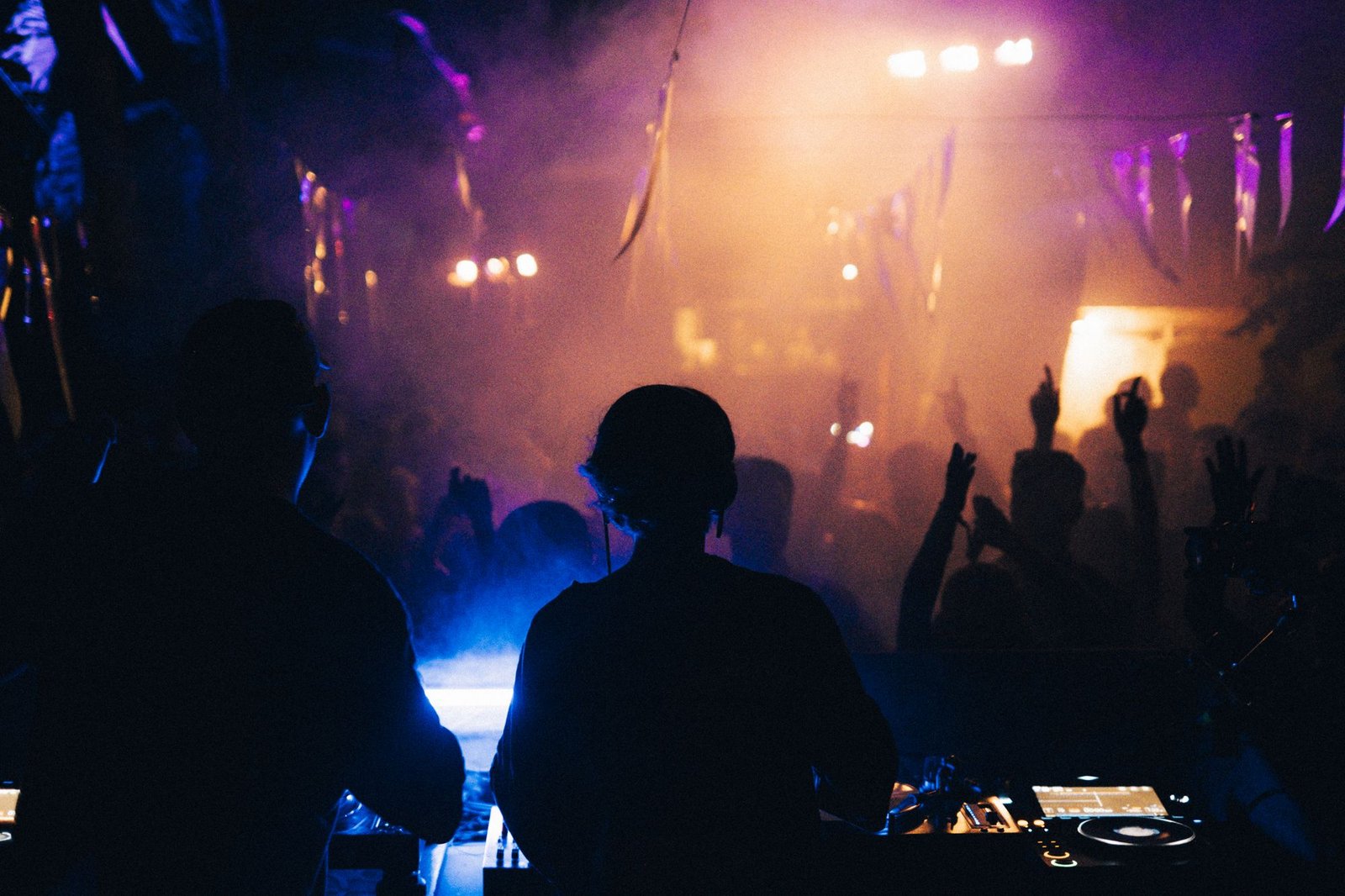 Two DJs at the decks, seen from behind, facing a crowd with hands up in purple and amber haze